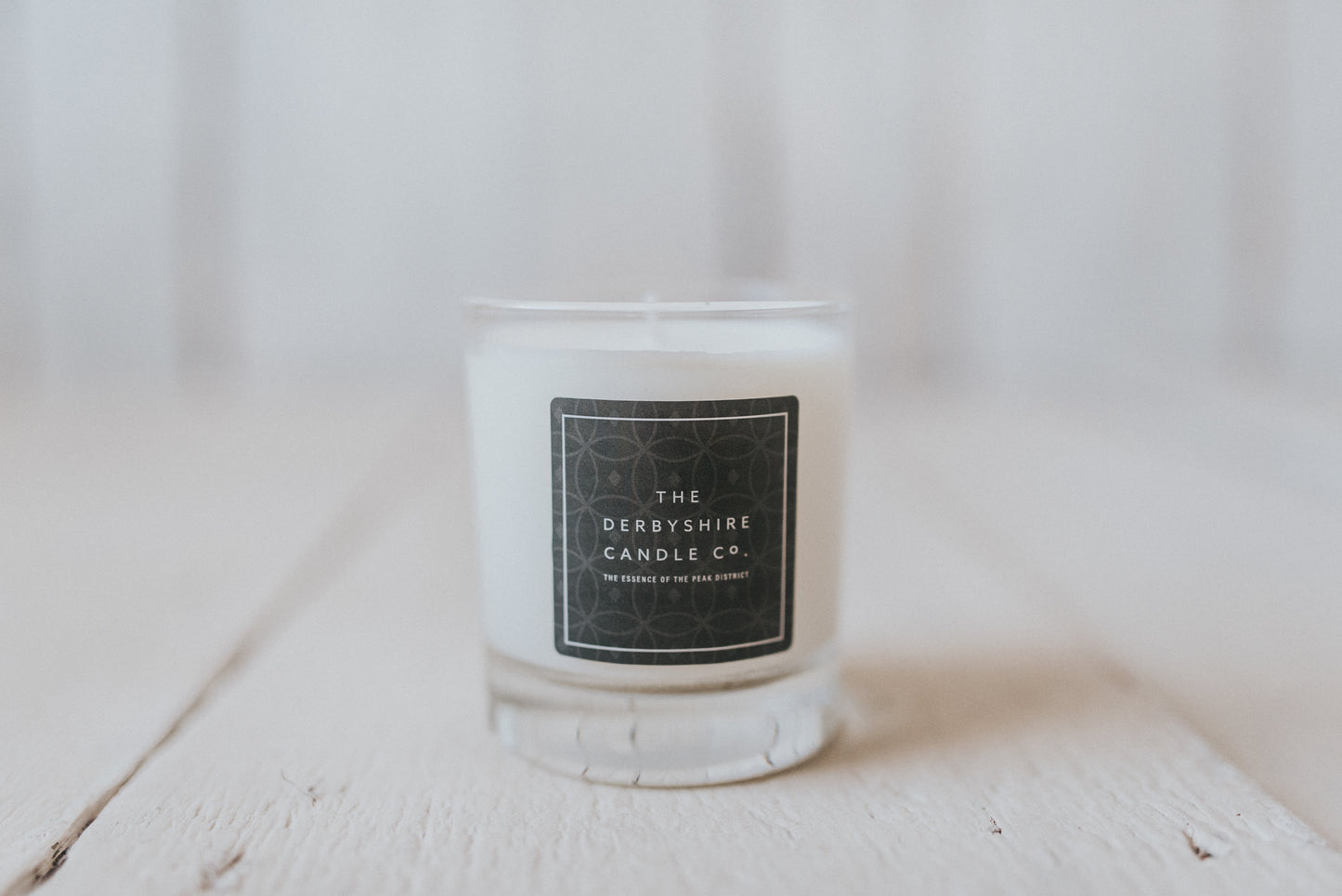 A white candle with a label that reads 'The Derbyshire Candle Company', housed in a clear glass holder, placed on a white wooden surface.